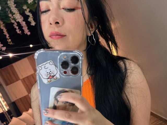 AriLynn, com cabelo preto, sombra rosa e brincos de argola, segurando um celular com um adesivo fofo de gato, tirando uma selfie no espelho com uma blusa laranja..