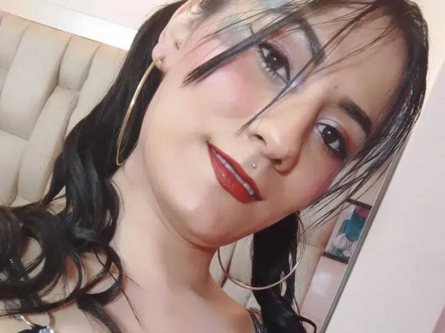 HotDellly has black hair in pigtails, red lipstick, a nose piercing, and big hoop earrings. Her makeup includes pink eyeshadow and a slight smile.