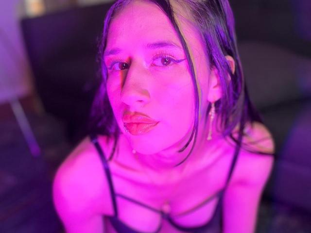 LilyRossi, avec un maquillage aux reflets violets, de longs cheveux lisses et de petites seins, portant un haut noir à bretelles, regardant vers le haut avec une légère moue, une lumière rosée-violette sur son visage..