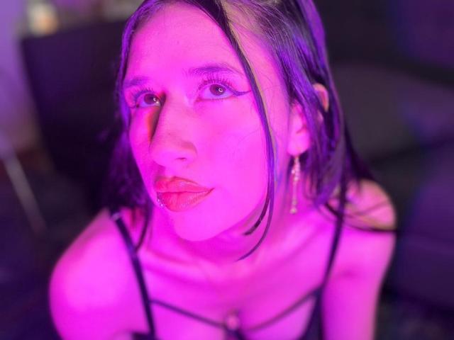 Lily Rossi mit einem rosafarbenen Teint, dunklem, glattem Haar, Eyeliner und rotem Lippenstift, trägt ein schwarzes Oberteil mit Trägern, Ohrringe und eine Halskette, im violetten Licht, und blickt nach oben..
