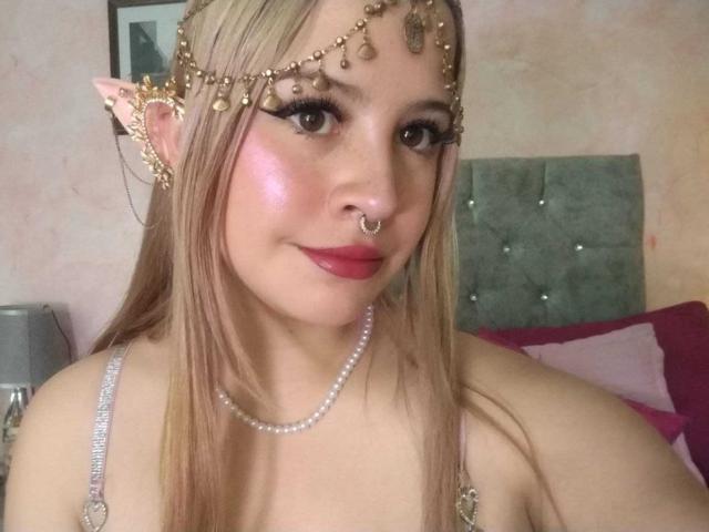 Melani Becker, con orejas puntiagudas, una diadema adornada con joyas, colorete rosa, lápiz labial rojo, un aro en la nariz, un collar de perlas y cabello rubio largo, mostrando sus pechos en un top plateado con tirantes..
