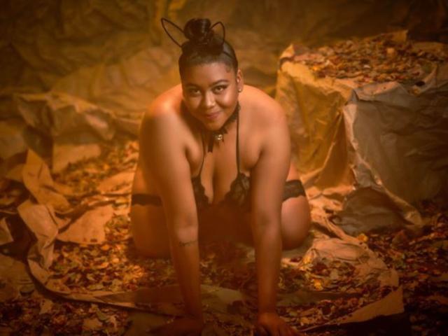 BiancaForest, crouching, wearing black lace lingerie with cat ears, smiling at camera, hands on ground, brown skin, black hair in bun, small tattoo on left arm, brown eyes.