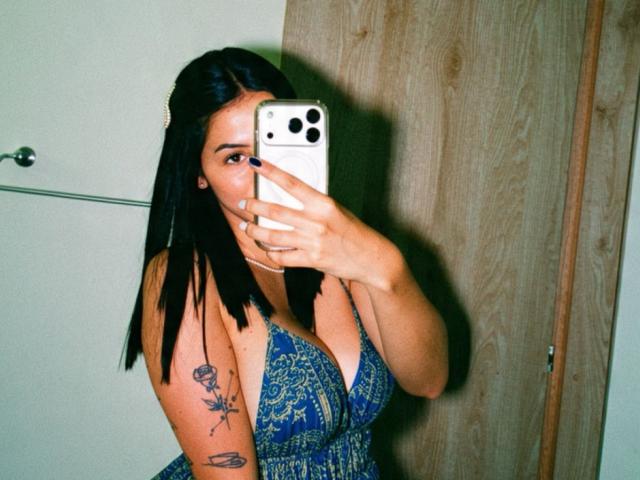 Isabella James mit langem, schwarzem Haar, einem blauen, gemusterten Kleid und Tätowierungen, macht ein Selfie mit einem weißen iPhone, auf dem ihre Brüste zu sehen sind, und hält das Telefon über ihren Mund..