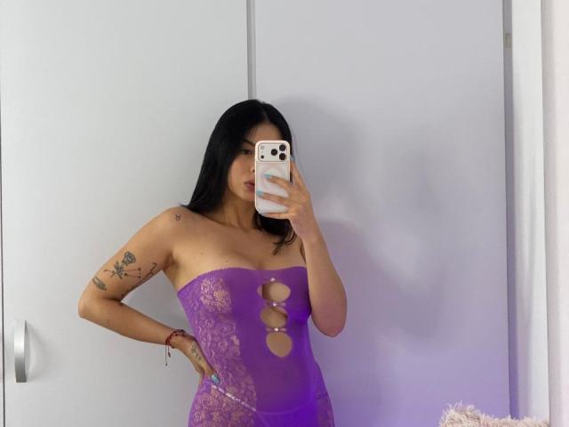 Isabella James mit langem, schwarzem Haar, trägt einen lila Overall mit Schnürung, der ihre Brüste zeigt, hat einen tätowierten Arm, hält ein Telefon und macht ein Selfie im Spiegel..