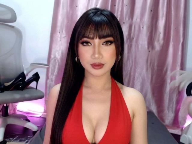TsLucyPearl with long black hair, bangs, and green eyes, wearing a red halter top that shows her big tits, standing in front of a pink curtain.