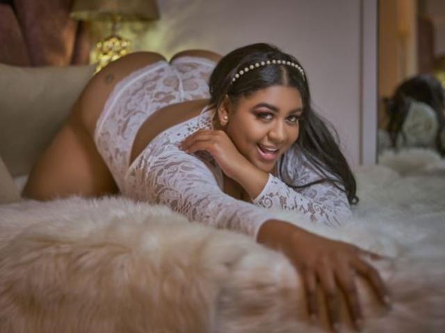 BiancaForest on all fours, wearing white lace underwear, smiling with her ass up, long black hair, pearl headband, on a fluffy white bed. Her tits are visible.