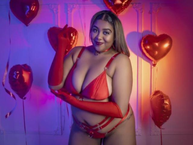 BiancaForest in red lingerie with long gloves, her big tits and curvy ass, standing with red heart balloons, smiling confidently, hands near face, in a colorful, purple-lit room.