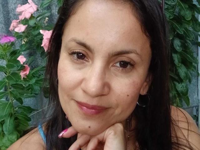 SolaraDubois with brown eyes, dark straight hair, pink lips, small nose, and a slight smile, resting her chin on her hand, wearing a blue top, and hoop earrings.