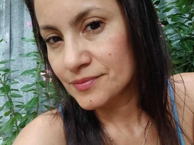 SolaraDubois has a cute face with dark brown hair, light pink lipstick, and a small beauty mark under her chin. She's smiling slightly with green plants in the background.