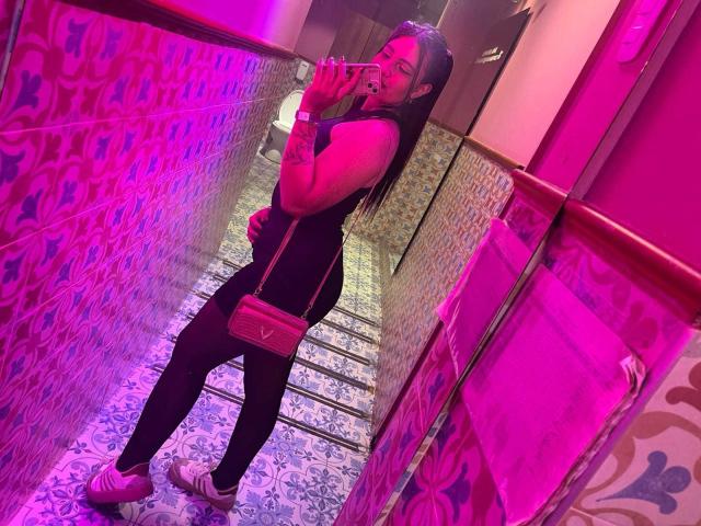 TammySmile with long black hair, wearing a black sleeveless top and black pants, holding a pink purse, taking a mirror selfie in a pink-lit bathroom, smiling.