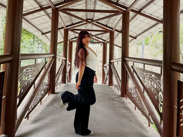 AlyssaGreen stands in a metal bridge, wearing a white tank top and black pants, lifting one leg, showing off her ass, with long brown hair and glasses.