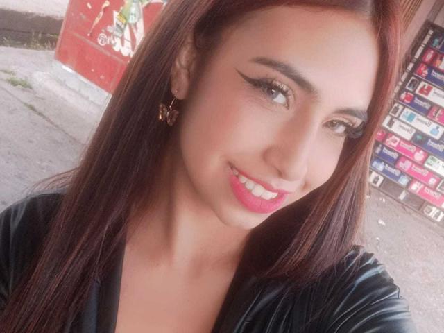 TammySmile has long brown hair, black eyeliner, pink lipstick, and a cute smile. She's wearing a black leather jacket and gold butterfly earrings. Her tits are not visible.