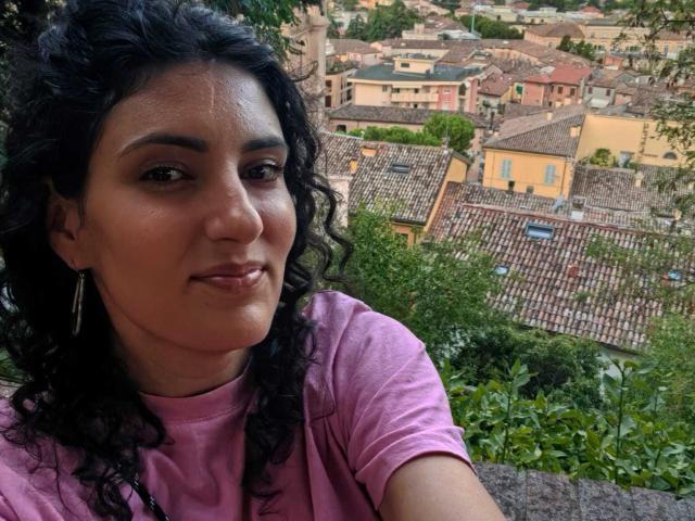 Curly-haired AddictedToUTS with medium brown skin and pink shirt, smiling at camera, showing small nose and subtle makeup, with greenery and rooftops in background.