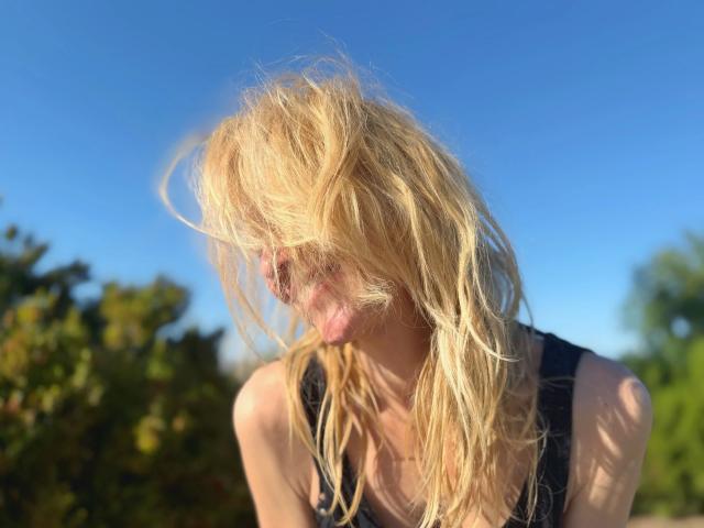 ShaiyaShy-hot with messy blonde hair, eyes squinting, tongue out, wearing a black tank top, blue sky background, sunlight highlighting her hair and face.