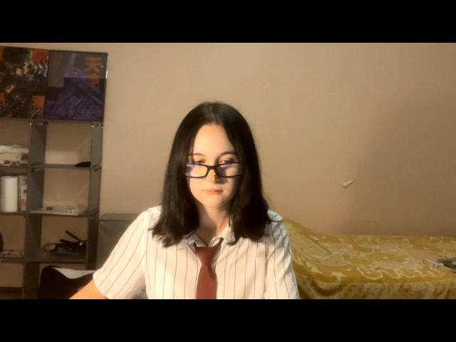 AustinKutz has medium-length black hair, glasses, and a white pinstriped shirt with a red tie. She's sitting, looking slightly to the side, with a yellow-patterned bed behind.