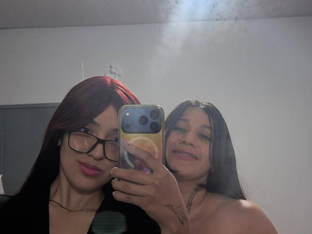 AbbyAndMichelross with red hair and glasses, and AbbyAndMichelross with black hair, both smiling, taking a selfie with a phone. AbbyAndMichelross on the right has a tattoo.