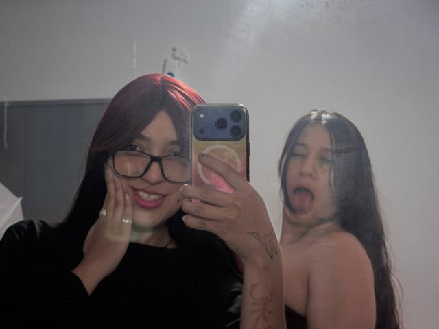 AbbyAndMichelross with red highlights, glasses, and black top, taking selfie. Michelross, topless, with long dark hair, tongue out, behind. Reflection, phone with pink grip, tattoo on hand.