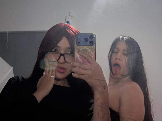 AbbyAndMichelross with red hair and glasses, holding phone, black top, hand on cheek, ring visible. Michelross with long black hair, tongue out, topless, tattoo on arm.