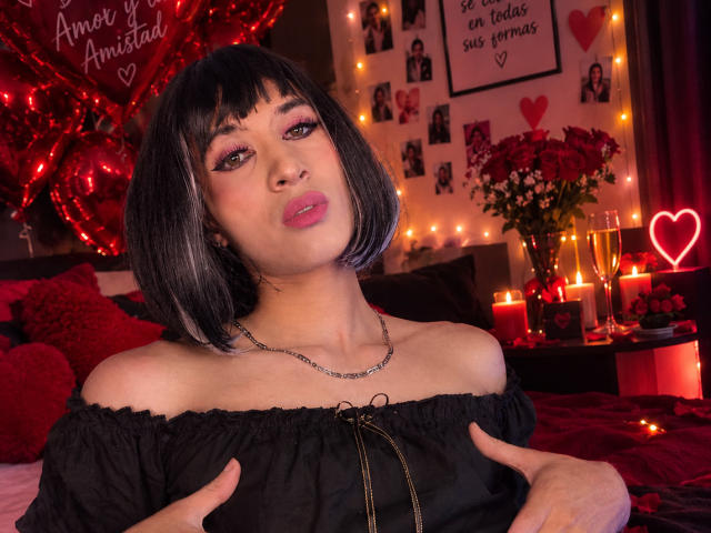 AlexiaHearts with black bob, pink eyeshadow, and red lipstick, wearing an off-shoulder black dress, hands on chest, necklace, surrounded by red candles and heart decorations, pouty expression.