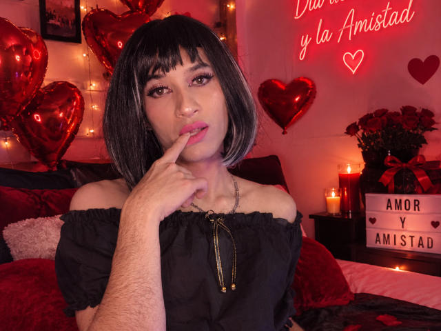 AlexiaHearts with a black bob, off-shoulder black top, finger to pink-lipped mouth, red heart balloons, candles, and neon heart on wall. She's got a cute, sexy look.