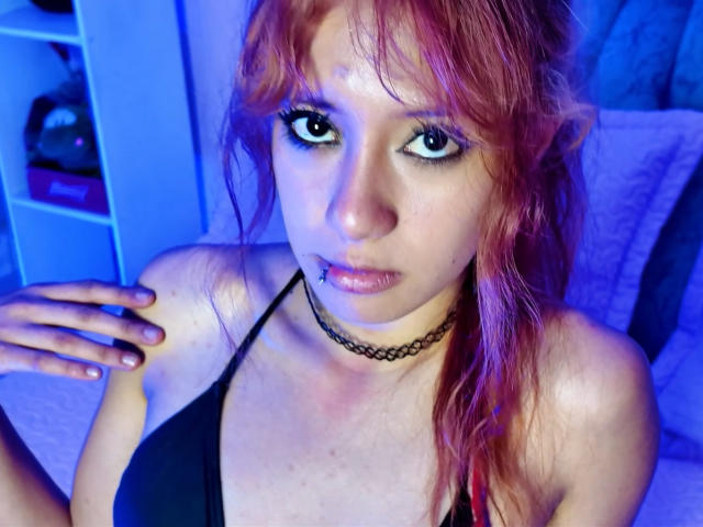SuriEverly with red hair, black choker, black top, and nose piercing, looking at camera with blue-tinted eyes, hand on shoulder, pinkish-red skin, slightly parted lips.