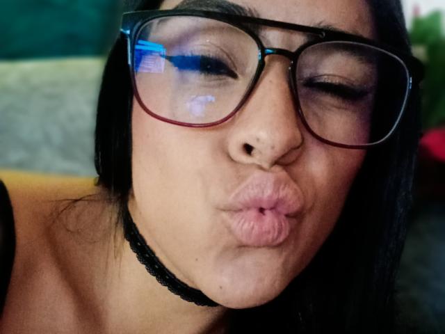 StellaBeckker's pouty face with large, red-rimmed glasses, black choker, and plump, pink lips, making a puckered expression. She has dark hair and a light skin tone.