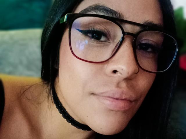 StellaBeckker, com um colar preto, usando óculos com armação vermelha, delineador azul e lábios levemente curvados, olhando para a câmera. Ela tem pele de tom médio e cabelo preto liso..