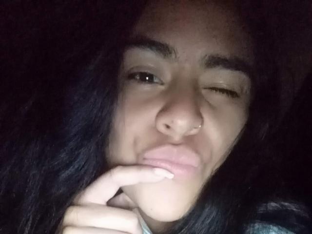 StellaBeckker with dark hair, winking, and touching her lips with her finger, nose pierced, in a low-light selfie. Her skin is tan, and she looks cute.