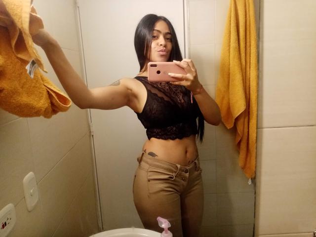 StellaBeckker with long black hair, wearing a black lace crop top, showing her tits, and tan pants, taking a mirror selfie with a pink phone in a bathroom.