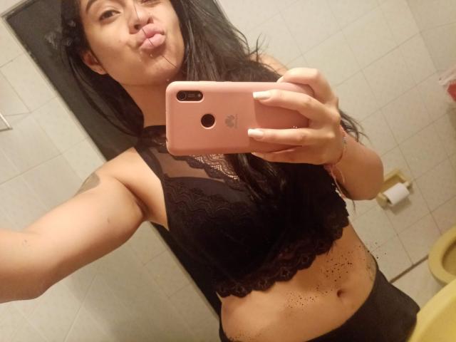 StellaBeckker with long black hair, puffy lips, black lace bra, holding pink iPhone, showing small tattoo on left arm, light skin, and slightly visible pubic hair.