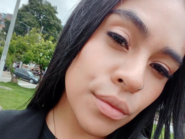 StellaBeckker's face with black hair, brown eyes, and full lips, wearing black eyeliner and a nose ring, smiling slightly, in a selfie with green grass in the background.