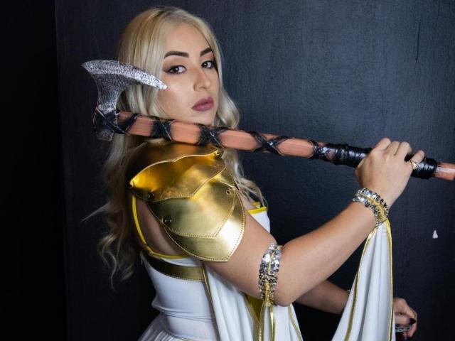 NashlyMilf avec des cheveux blonds, une armure dorée et une robe blanche, tenant une hache à double tranchant, regardant par-dessus son épaule, portant un bracelet en argent, dans un décor sombre, mettant en valeur ses seins..