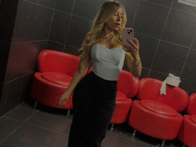 AmaraRicci with blonde hair, wearing a white strapless top, black skirt, and choker, taking a mirror selfie in a room with red leather chairs. Boobs visible under top.