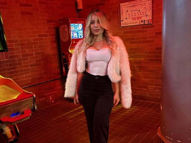 AmaraRicci with blonde hair, white fur jacket, white strapless top, black high-waisted pants, and a pearl necklace, standing in a red-lit brick room, smiling.