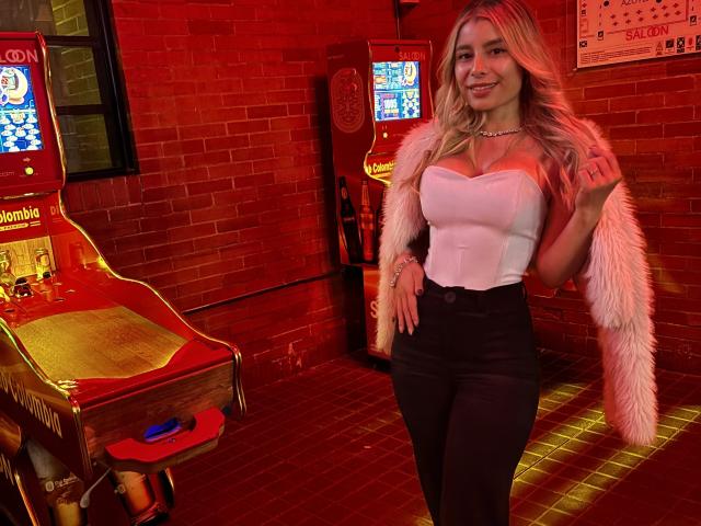 AmaraRicci with blonde hair, white strapless top, black pants, and pink fur coat, standing by red-lit arcade machines, smiling, showing her tits. Her ass is covered.