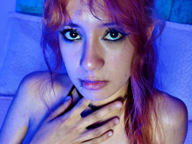 SuriEverly with red hair, blue eyes, and a nose piercing, holding her chest with her hand, wearing black eyeliner, and a black choker, looking up with a slightly parted mouth.