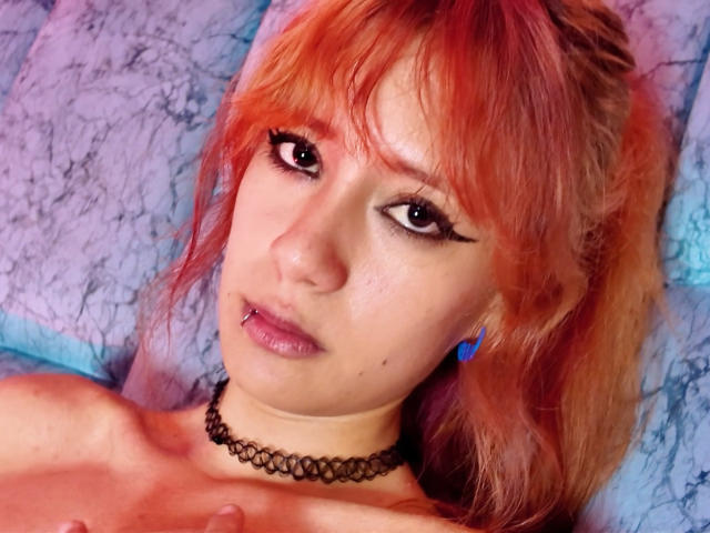SuriEverly has red-orange hair, black eyeliner, purple lipstick, a black choker, and blue earrings. Her topless chest is slightly visible with her hand near her collarbone.