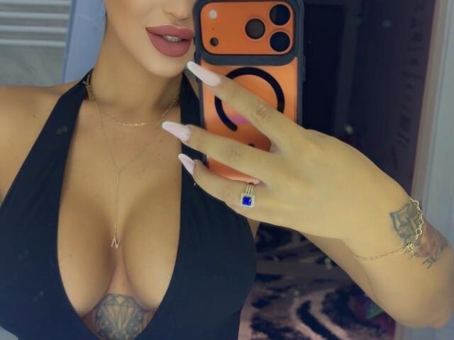 NicolSNaya's big tits in a black halter top, holding an orange phone with a ring, long nails, tattoo on her wrist, and a blue ring, with red lipstick.