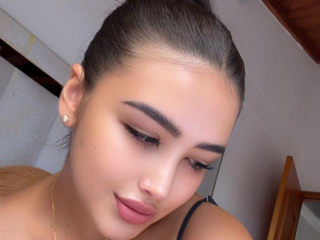 AnnaPooky's face with smooth, tan skin, dark eyebrows, long eyelashes, and pink lips. She's looking down with a slight smile, wearing a black strap and gold earring.