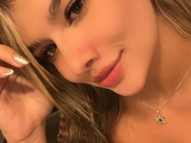 SashaPolet's face with light brown skin, blonde hair, and subtle smile. She's wearing a sunflower necklace, has glittery nails, and a slightly tilted head. Her tits are not visible.