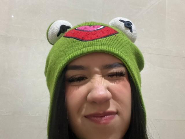 HannaThorne with green frog hat, eyes closed, smiling slightly, nose small and round, dark hair, cute, white eyeball-like hat decorations, pink mouth, soft skin, and small black nose.