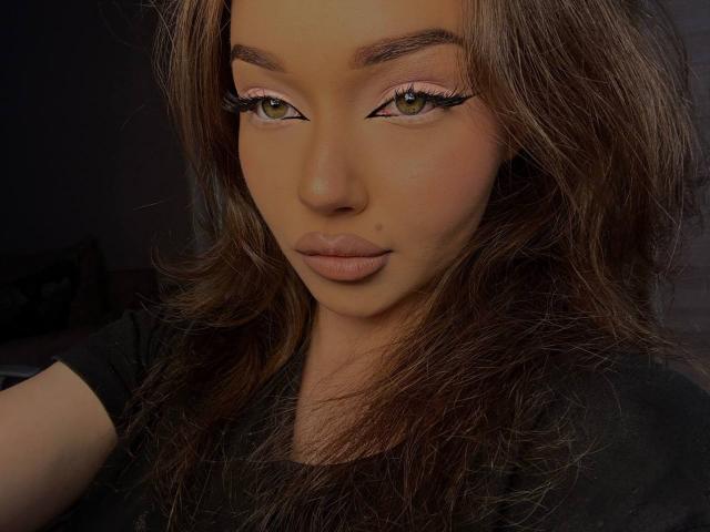 GretchenJamili has long brown hair, brown eyes with black eyeliner, pink eyeshadow, and full pink lips. She's wearing a black shirt, looking serious and sexy.