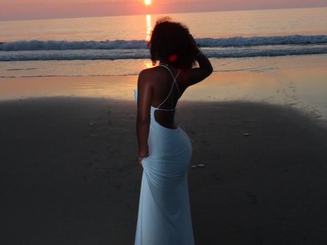 CelesteCharme stands on a beach at sunset, wearing a white, backless dress, with curly hair, hand in dress, and her ass facing the camera, reflecting the orange sky.