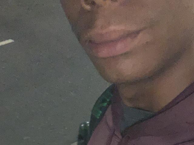 AndyCris's lower face with slightly parted pink lips and a small goatee, wearing a purple shirt, has a dark skin tone and is photographed in a dimly lit setting.
