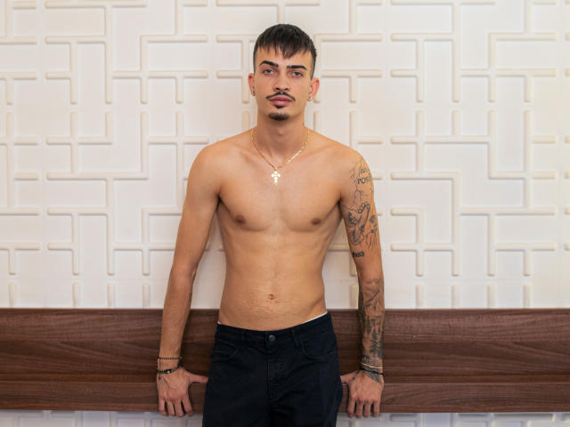 Brandon Tate sem camisa, com um braço tatuado, bigode e colar, apoiado em um banco de madeira, vestindo calças pretas e olhando para a câmera. Suas nádegas não estão visíveis..