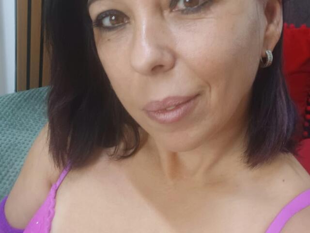 LauraSin has purple hair, brown eyes, and is wearing a pink top with visible cleavage. She's smiling slightly with a subtle nose ring and earrings.