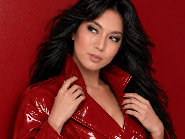 Chanteuse with wavy black hair, red glossy leather jacket, and subtle makeup, holding collar, looking off-camera, showing small tits and a sexy, confident expression. Red background.