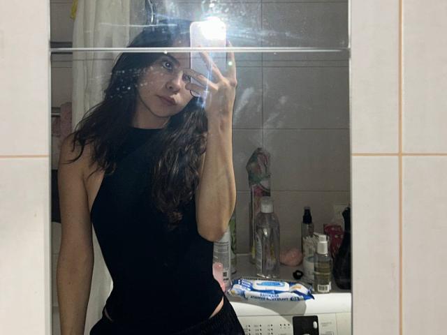 AnneKofron with wavy dark hair, wearing a black sleeveless top, taking a mirror selfie showing her bare ass and black underwear, holding a phone with a bright flash.