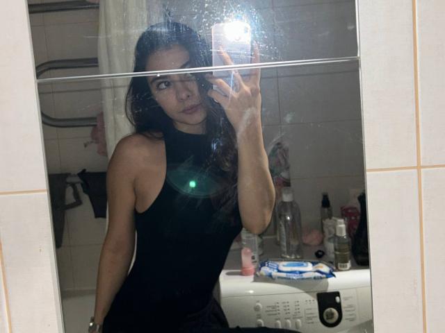 AnneKofron with long dark hair, wearing a black sleeveless top, taking a mirror selfie, showing her small, perky tits and flat ass, in a bathroom with a washing machine.