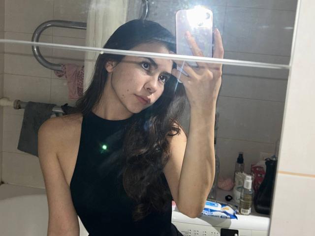 AnneKofron with long brown hair, black sleeveless top, taking a mirror selfie in a bathroom, showing her tits and a small mole on her cheek, holding a phone.
